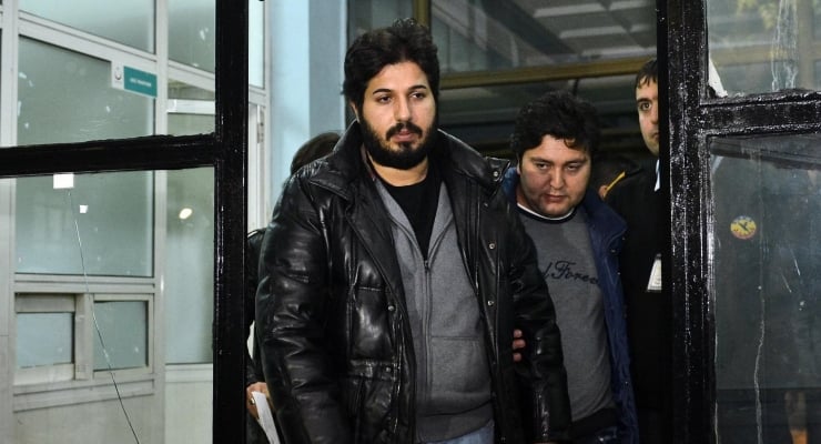 Bearded man in a black leather jacket walks through a glass door, flanked by two men indoors.