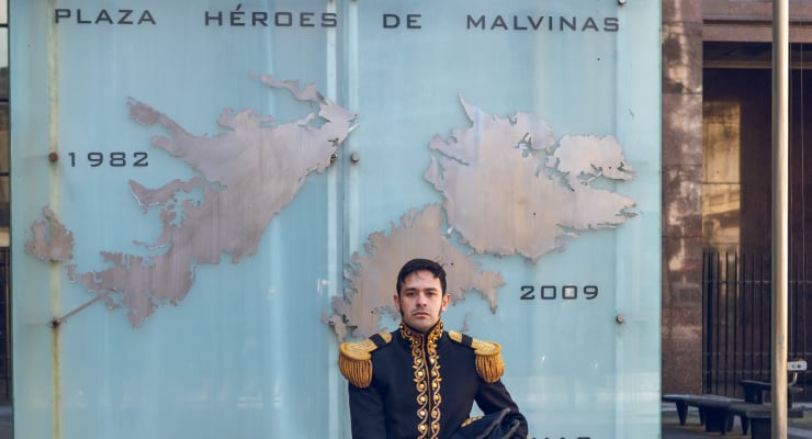 Man in an ornate ceremonial uniform standing in front of a turquoise commemorative wall featuring a metal map of the Falkland Islands and the years 1982 and 2009, titled Plaza Héroes de Malvinas (Falklands Heroes Square).