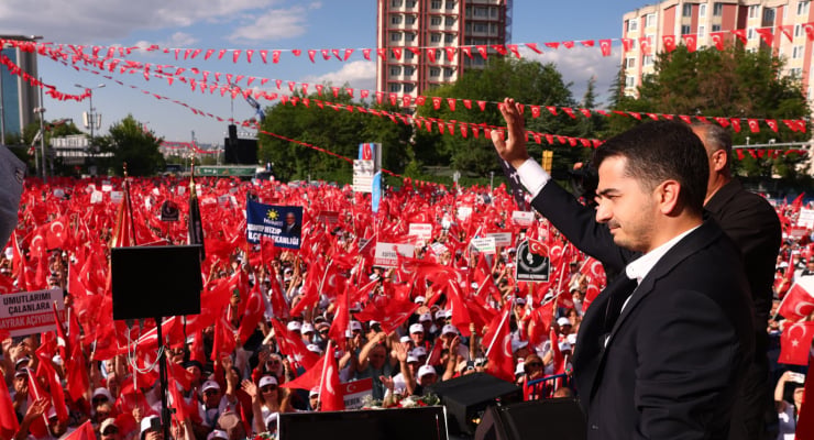 Politician in a dark suit at a podium, raising his hand to a large outdoor crowd waving red flags at a political rally.