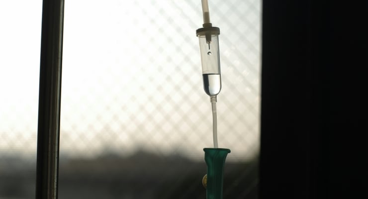 IV drip bag with tubing hanging from a stand in a dim room, ready for infusion.