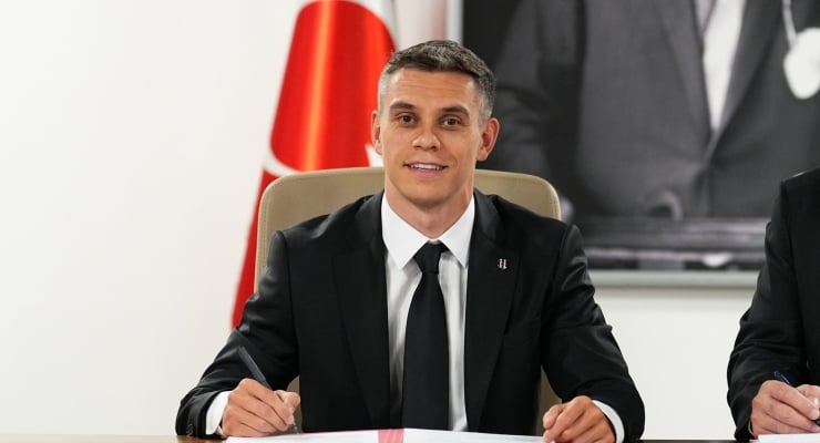 Man in a black suit and tie signs a document at a table, with a Turkish flag in the background.