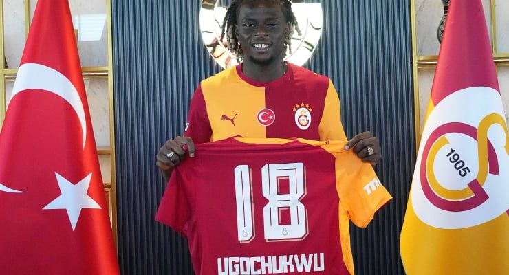 Smiling footballer in Galatasaray colors holds up a red and yellow jersey with number 18 and the name UGOCCHUKWU, standing between a Turkish flag and a team flag.