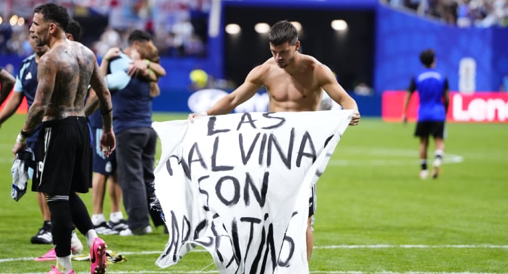 Two shirtless soccer players on a green pitch hold up a white banner with black letters; other players and spectators are in the background.