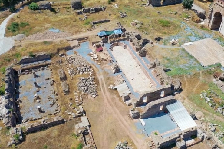 Aerial view of ancient ruins with a rectangular central arena, stone walls, and scattered stones across the site.