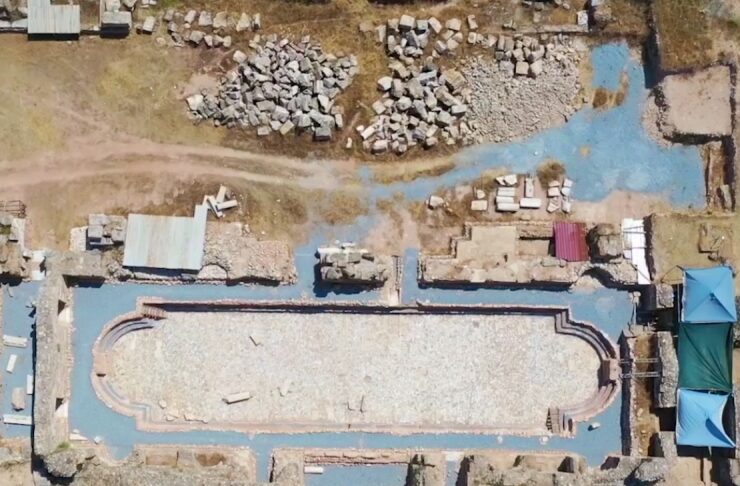 Aerial view of an ancient ruin featuring a rectangular stone pool with rounded ends, surrounded by stone walls and scattered rubble, with blue ground coverings and tents to the right.
