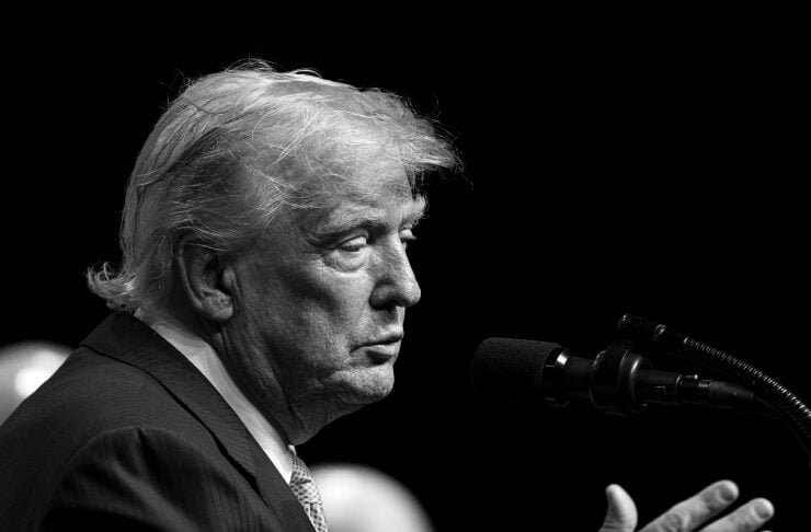 Black-and-white photo of an older man in a suit speaking into a microphone at a podium, gesturing with his hand on stage