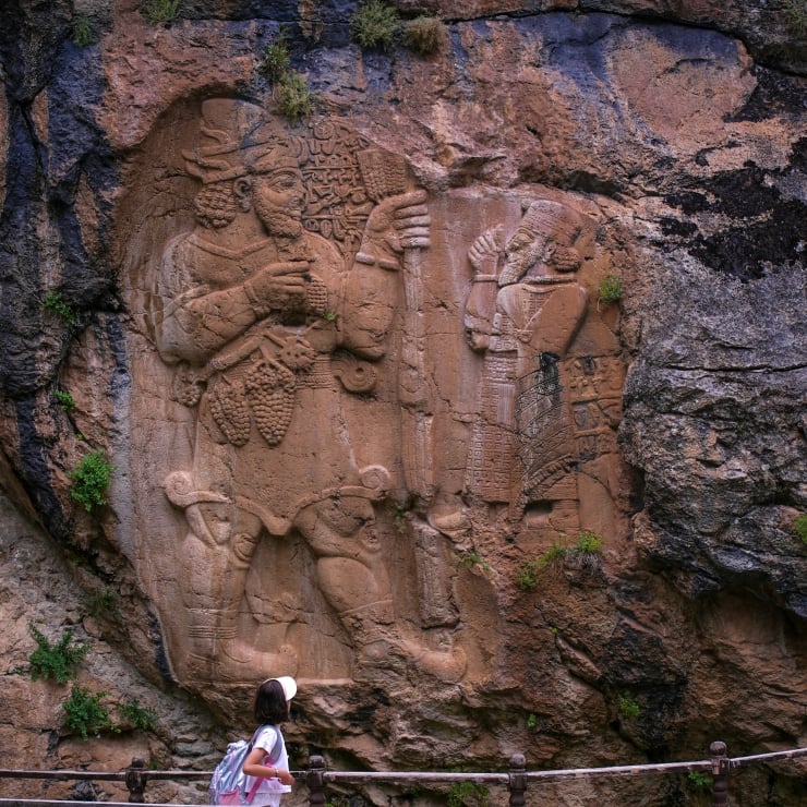 Bas-relief of two ancient warriors carved into a rock wall, with a girl and railing in the foreground.