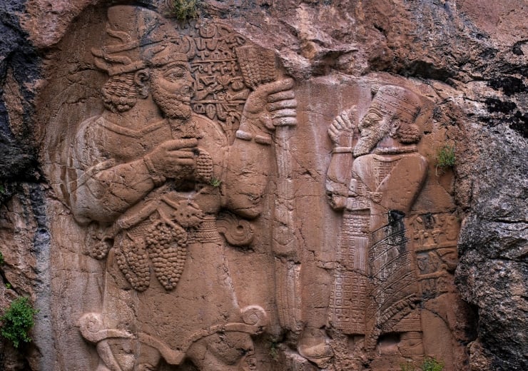 Ancient stone relief in a rock alcove showing two bearded figures in ceremonial attire, exchanging or presenting objects.