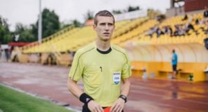 Referee in a yellow FIFA badge shirt stands on a running track at a stadium, holding a red card.