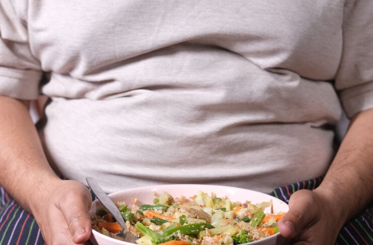 Person in a light shirt and striped pants holds a pink bowl of mixed vegetables and grains with a spoon.