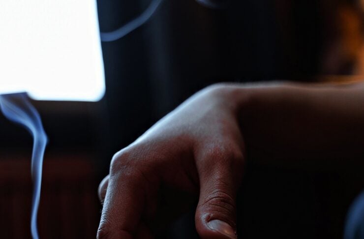 Close-up of a hand holding a lit cigarette, smoke curling upward in a dim room near a window.