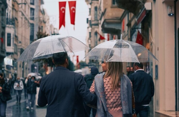 Couple walking on a rain-soaked city street under clear umbrellas, Turkish flags hanging above.
