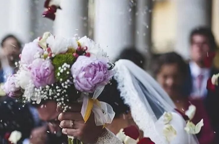 Bride in a white lace dress lifts a pink-and-white bouquet as petals float in the air, with a veil trailing behind.