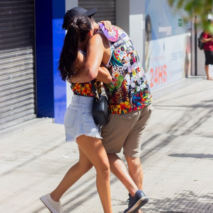 Two people share a hug on a sunny sidewalk, wearing colorful summer outfits.
