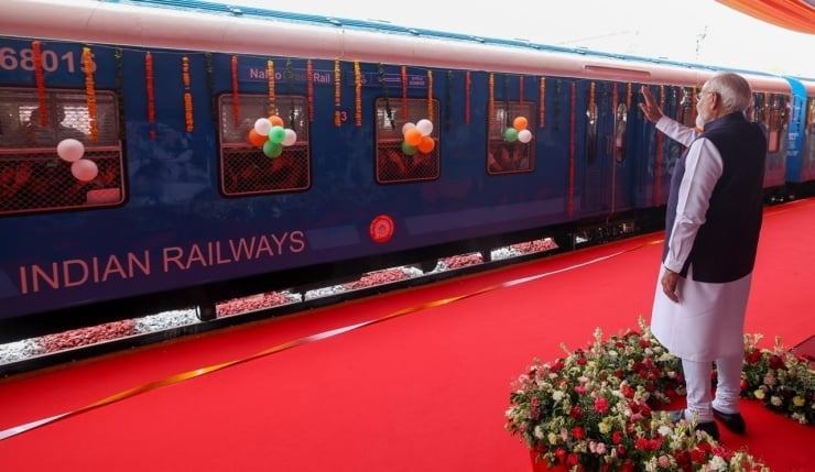 Blue Indian Railways train decorated with balloons on a red carpet; a man in traditional attire waves at the event