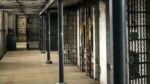 Prison corridor with metal bars and locked cell doors along a dim concrete hallway.