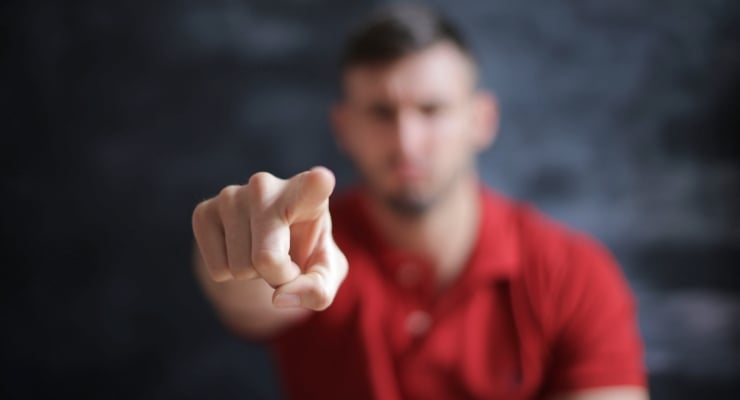 Blurred man in a red shirt points directly at the camera, addressing the viewer.