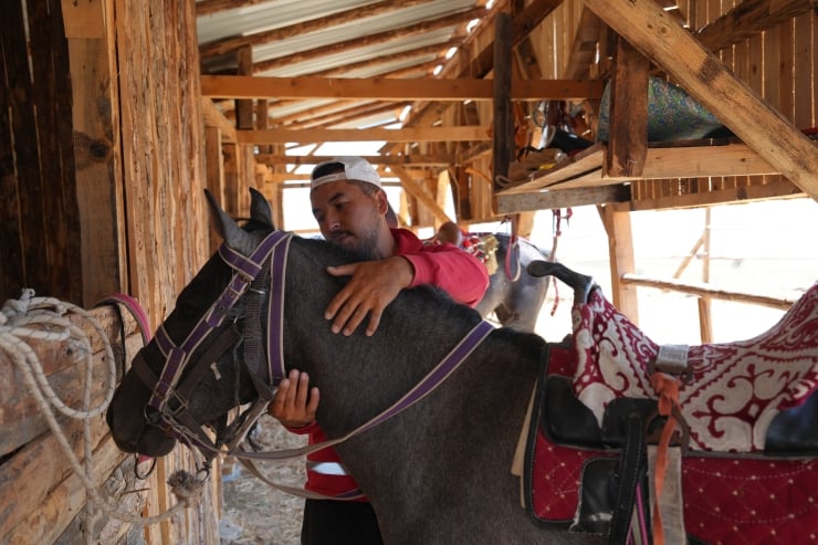Man in a red shirt and white cap hugs a gray horse inside a wooden stable, saddle and reins visible on the horse.