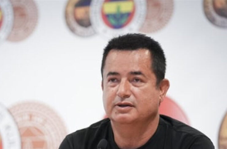 Man in a black t-shirt speaks into a microphone at a press conference, seals on the backdrop behind him.