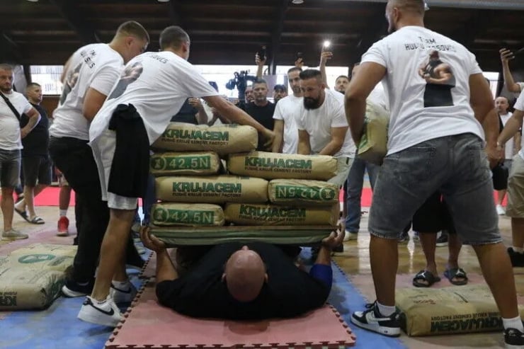 Group of men in white shirts participate in a strongman event, lifting stacked cement bags off a man lying facedown on a mat while onlookers photograph and cheer.