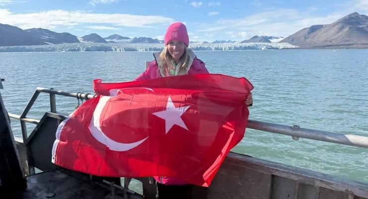 Woman in a pink hat and jacket on a dock, holding a Turkish flag with a glacier and mountains in the background.