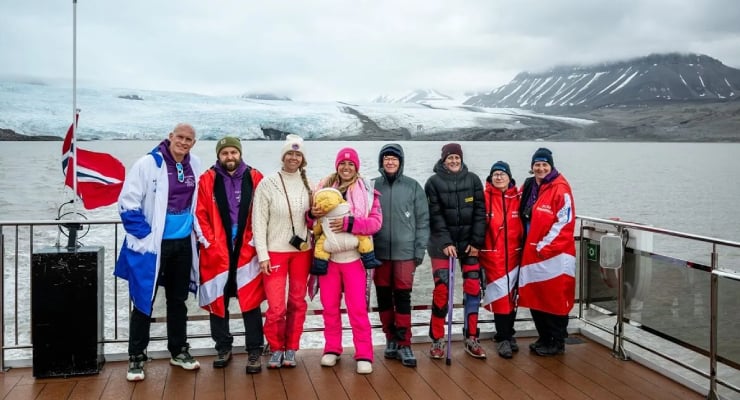 Group of eight adults and a child on a ship deck in cold weather, wearing bright jackets; Norwegian flag and snowy mountains in the background.