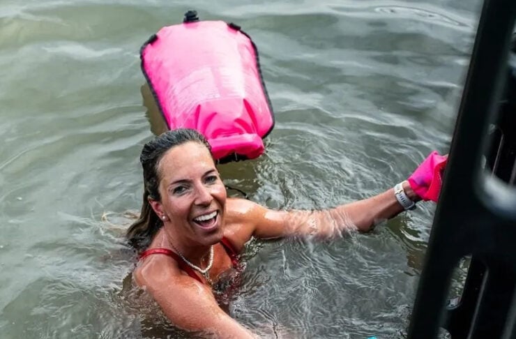 Smiling woman in open water wearing a red swimsuit; a pink life jacket floats behind her as she reaches for a white snorkeling mask near a boat ladder.