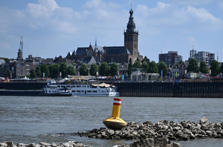 Yellow buoy resting on a rocky jetty by a river with a city skyline and tall church tower in the distance, under a blue sky.