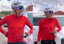 Cyclist in a red jacket and white helmet chats beside a white car, gloved hands on hips, wearing blue sunglasses.