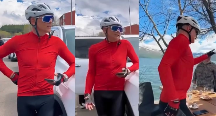 Cyclist in a red jacket, white helmet and blue sunglasses stands beside a car with hands on hips.