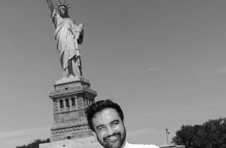 Man in a white shirt and tie posing with arms crossed on a railing, Statue of Liberty in the background.