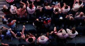 Aerial view of a crowded stadium stands with fans wearing red, white, and blue, many raising their hands in celebration.
