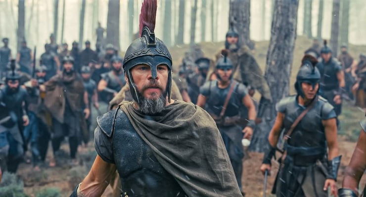 Leader in a Roman-style helmet with a red plume leads a group of armored soldiers through a forest clearing, cape draped over his shoulder.