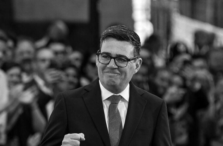 Man in glasses and a dark suit gives a fist-pump gesture at a public event, crowd blurred in background.