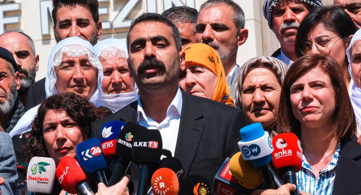 Central man in a dark suit speaks to reporters as a crowd of people surrounds him, microphones from many news outlets held toward the speaker.