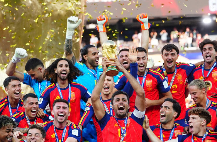 A men's soccer team in red and blue uniforms celebrates a victory, lifting a trophy amid gold confetti and cheering players.