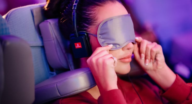 Passenger wearing a sleep mask and headphones while seated on an airplane seat in a dim cabin lighted scene, relaxing.