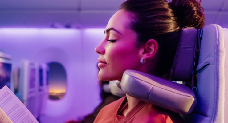 Profile of a woman with eyes closed, resting against an airplane seat headrest in purple cabin lighting while reading a document