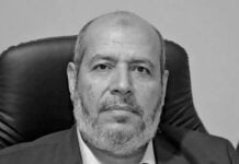 Middle-aged man with a gray beard in a dark suit and white shirt, seated in a black leather chair, facing the camera.