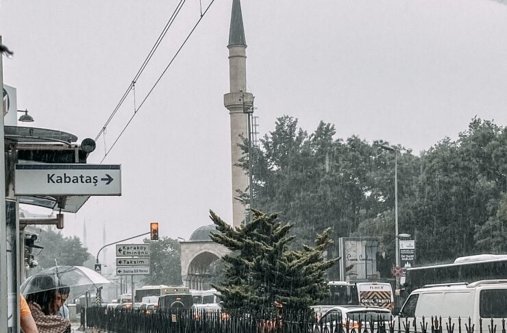 Rainy street in Kabataş, Istanbul: pedestrians with umbrellas at a station, a mosque minaret in the distance, and traffic on a wet road with a fenced median.