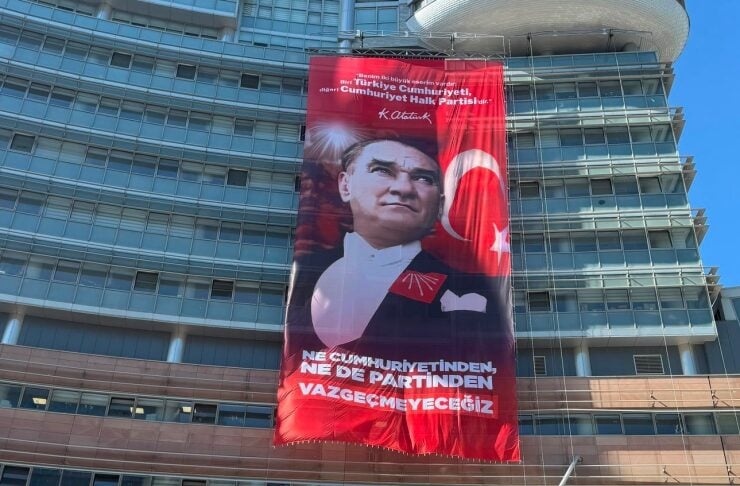 Large red banner with a portrait of Atatürk and the Turkish flag hanging from a modern glass building façade; Turkish text readable on banner and building sign nearby.