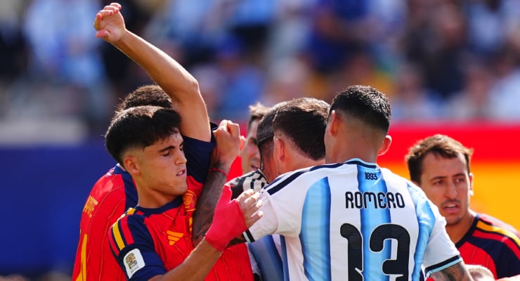 Soccer players from opposing teams grapple and push each other during a heated moment on the field, with a blurred crowd in the background.