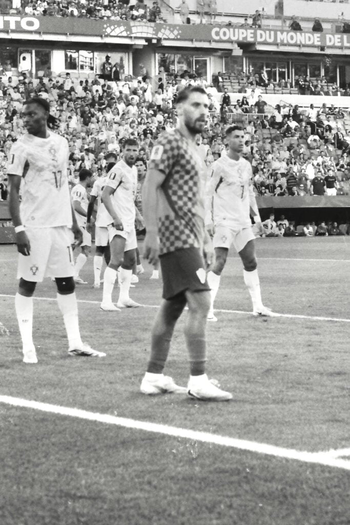 Soccer players in white uniforms on the field before a match, with a crowded stadium in the background and a banner visible above the stands.