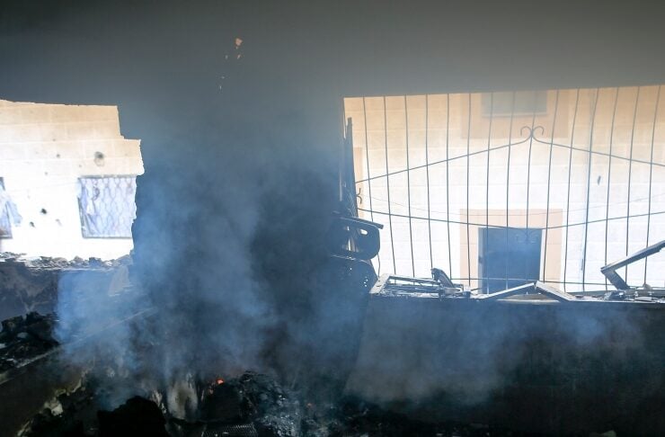 Charred rubble and smoke inside a burned room, with a metal gate and daylight visible through a broken window.