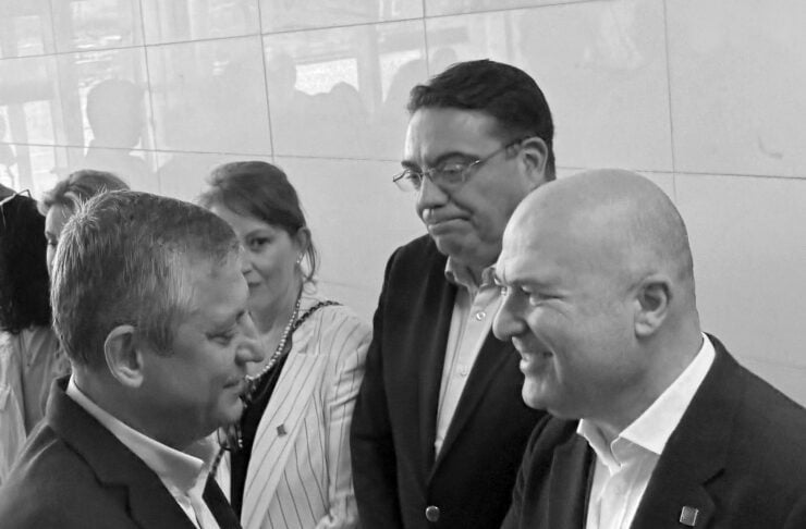 Two men in suits shake hands and smile in a professional greeting, with others watching in the background.