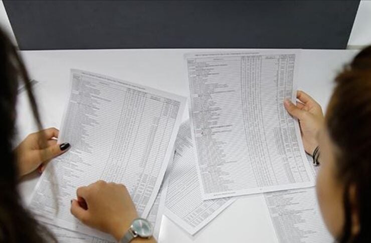 Overhead view of two people at a white table examining several printed spreadsheets or lists on the desk.