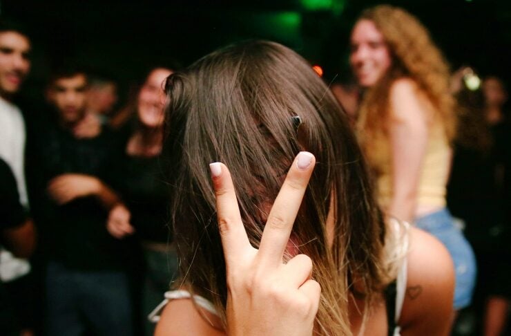 Person with long hair making a peace sign in front of their face at a crowded party or club.