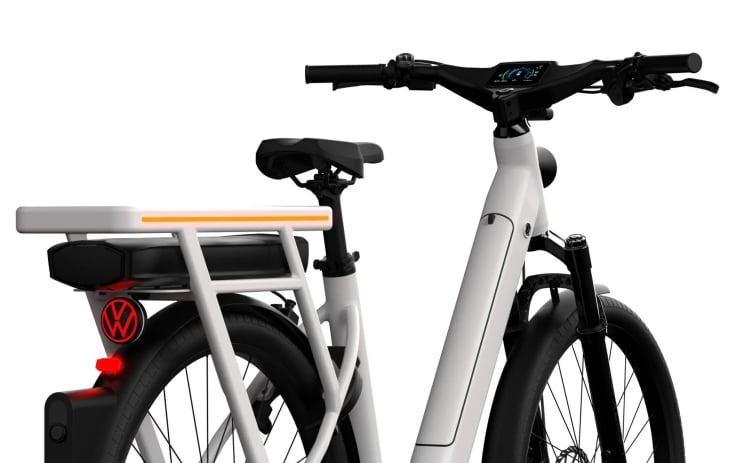 White electric bicycle with a front-display on the handlebar, thick frame, and rear cargo rack with an orange safety strip and light.