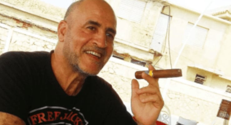 Middle-aged man with a shaved head and beard smiles while holding a cigar outdoors.