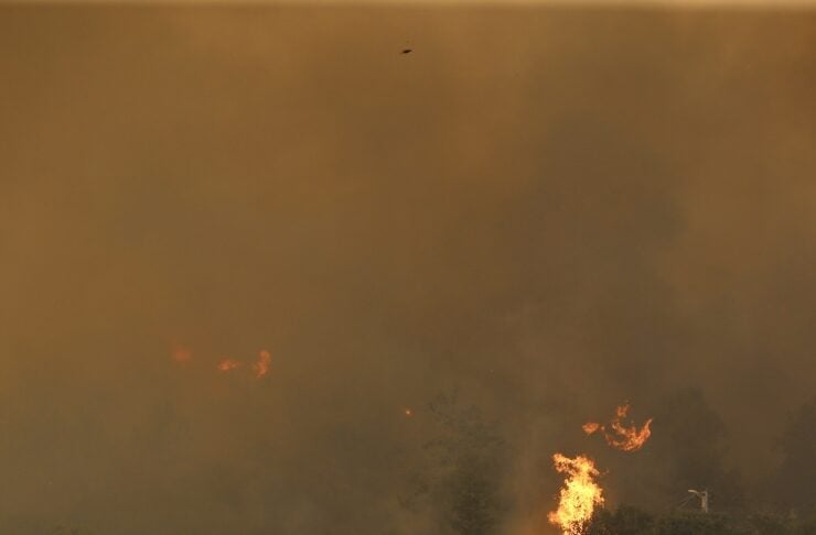 Wildfire near a hillside home with tall flames in trees and heavy smoke filling the scene.